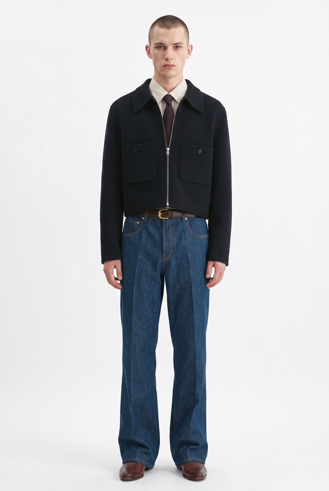 Man in a navy cropped jacket, tie and wide indigo jeans with brown shoes